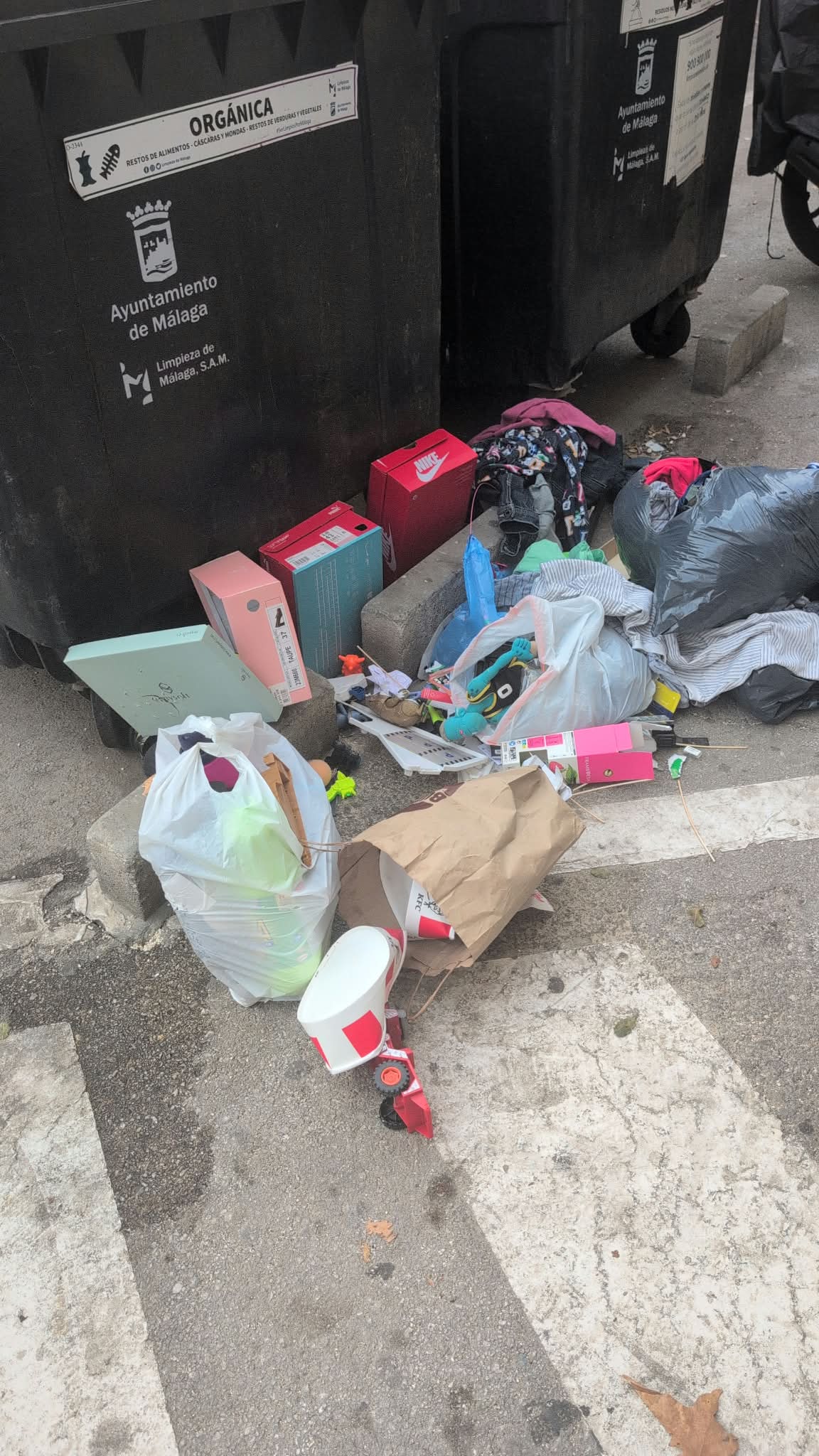 Bolsas, cajas de zapatillas y ropa junto a un contenedor del Ayuntamiento de Málaga, con un peluche azul asomando